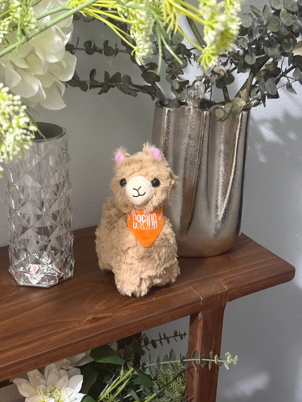 Plush toy llama with an orange bandana on a wooden surface with decorative vases and plants.