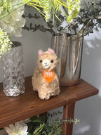 Plush toy llama with an orange bandana on a wooden surface with decorative vases and plants.